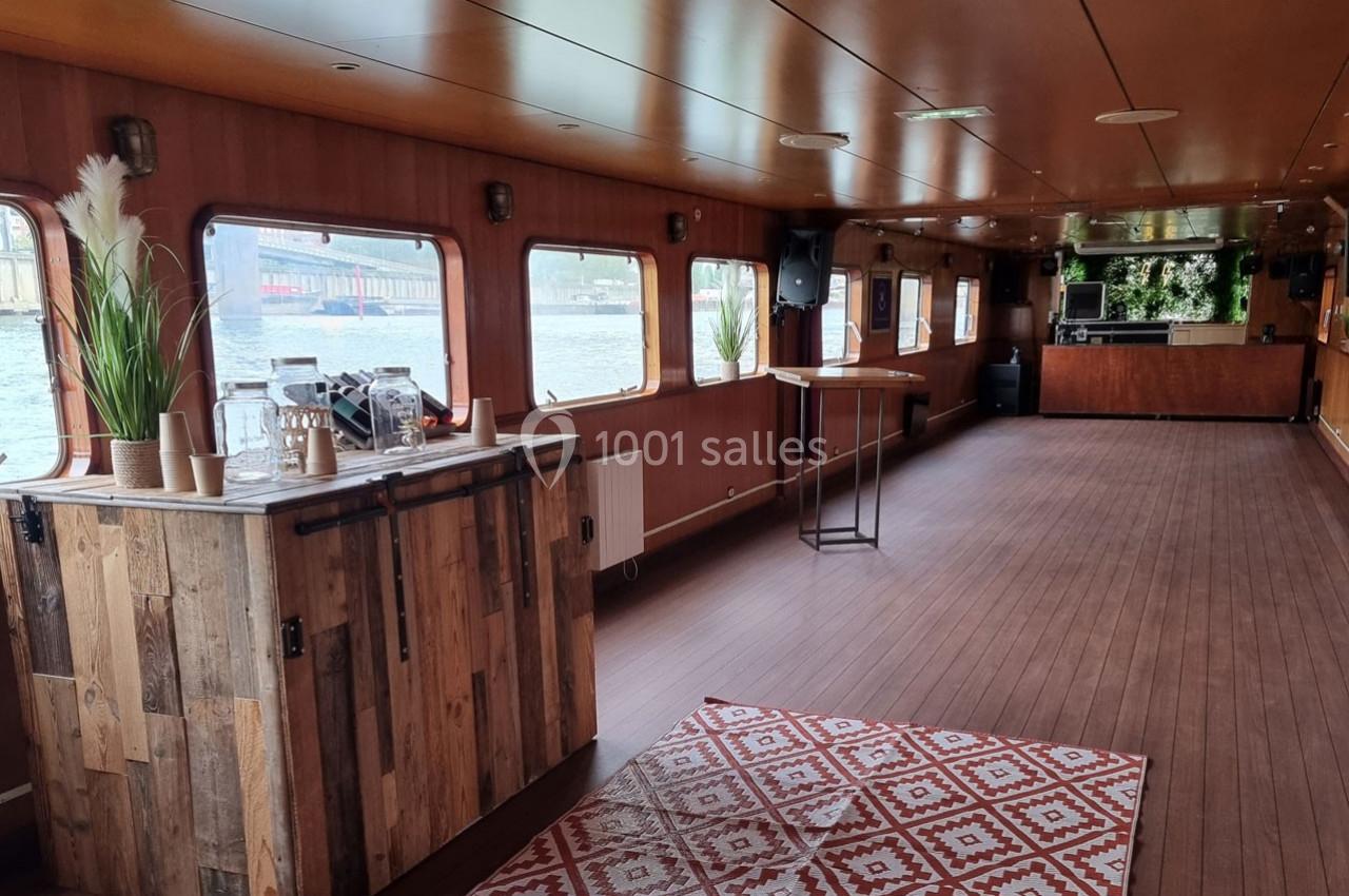 Intérieur d'une péniche avec parquet, bar en bois, grandes fenêtres et décoration végétale.