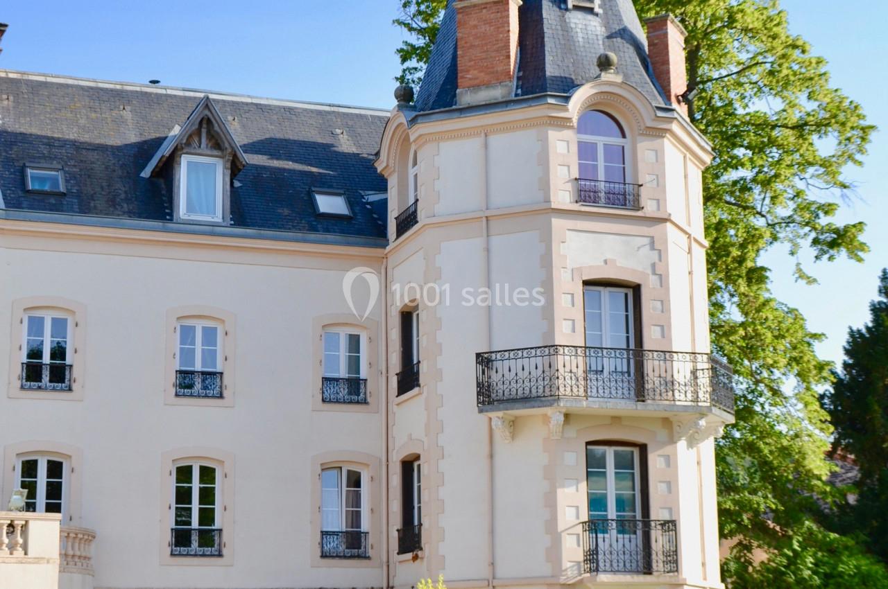 Façade d'un bâtiment ancien avec une tour, des balcons et des fenêtres, entouré de végétation et d'un parterre fleuri.