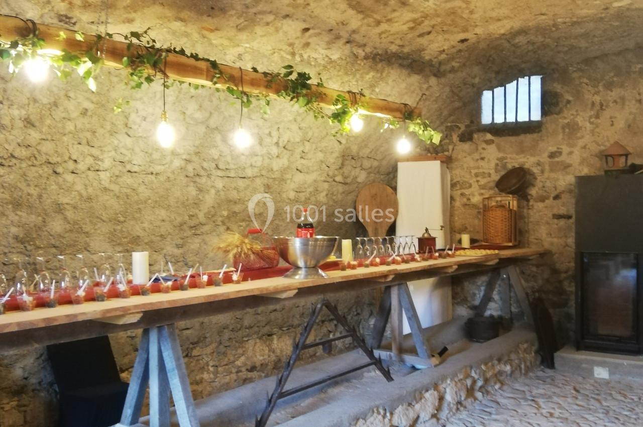 Table en bois avec verres et bouteilles alignés dans une cave voûtée en pierre, éclairée par des guirlandes lumineuses.