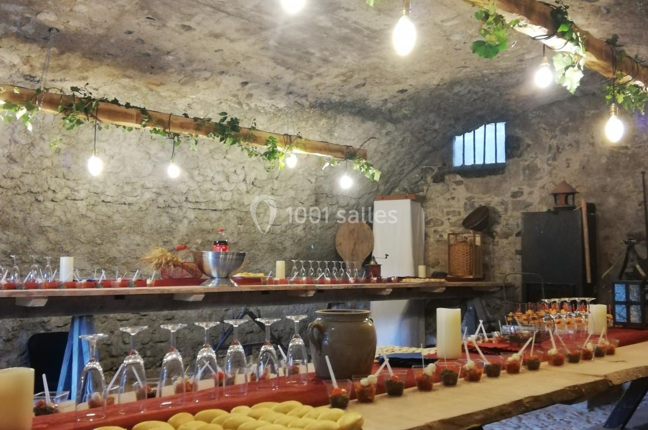 Salle voûtée en pierre avec une longue table en bois garnie de verrines, plats et décorations lumineuses.