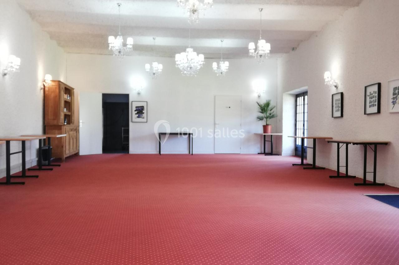 Salle spacieuse avec moquette rouge, murs blancs, lustres suspendus et tables alignées le long des murs.