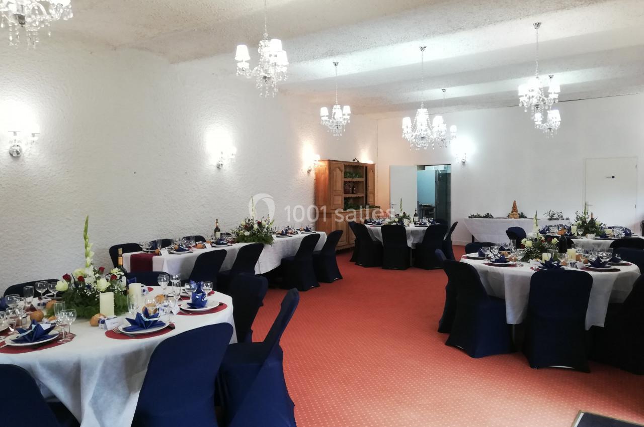 Salle de réception décorée avec des tables rondes dressées, nappes blanches, chaises bleues et lustres suspendus.