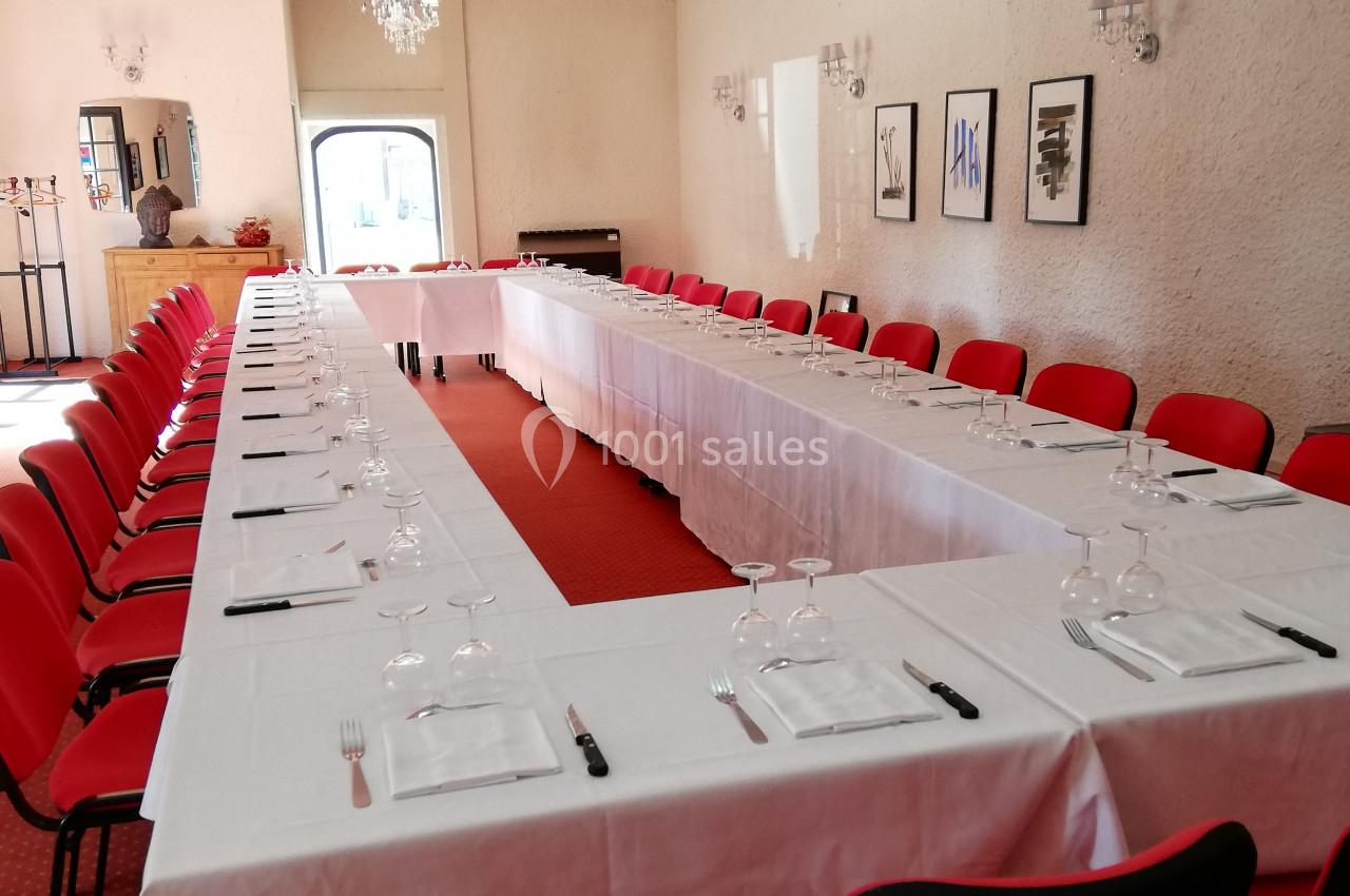 Salle de réunion avec tables en U couvertes de nappes blanches, chaises rouges et verres disposés pour un événement.