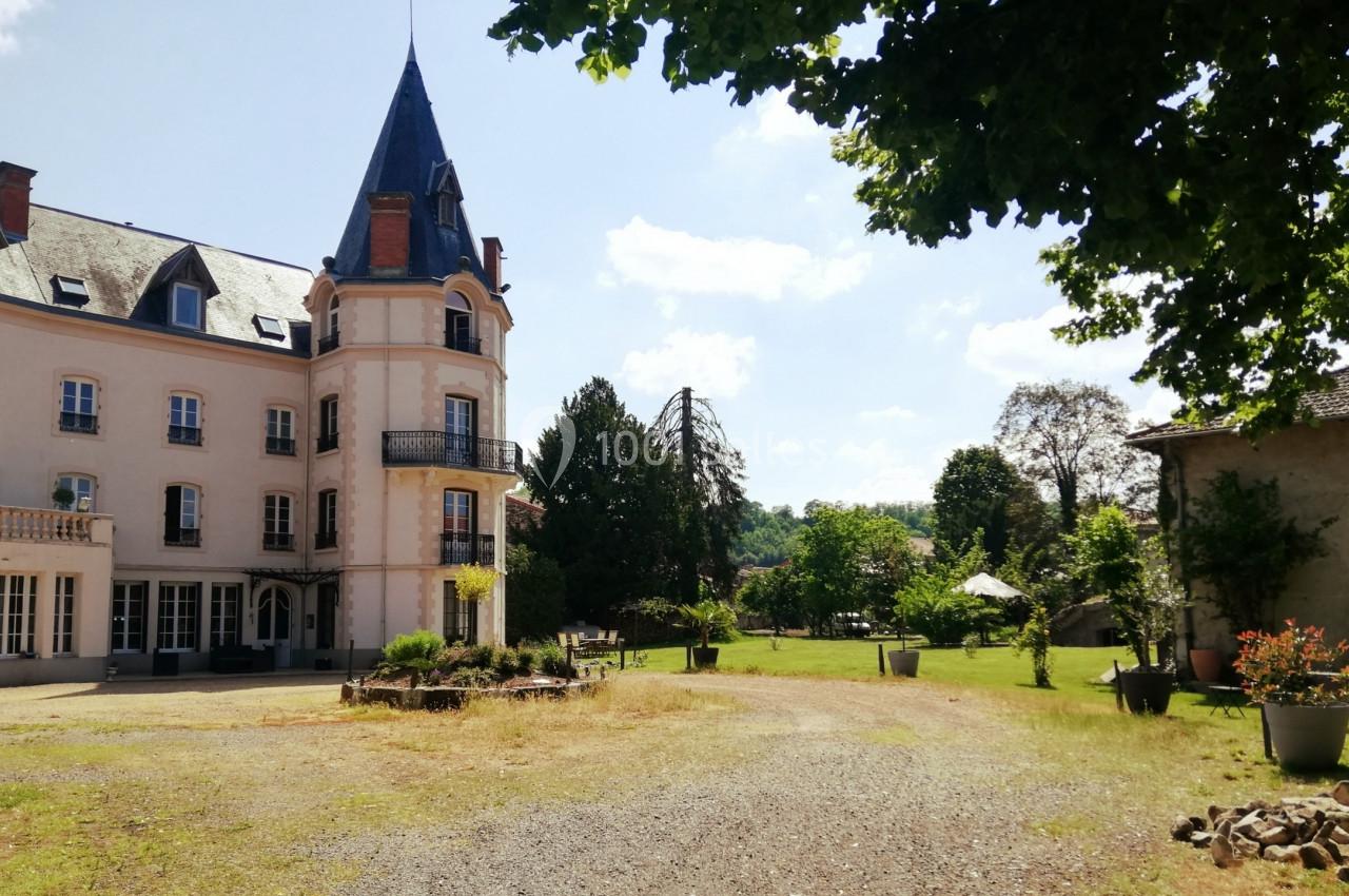 Façade d'un château avec une tour, entouré d'un jardin arboré sous un ciel ensoleillé.