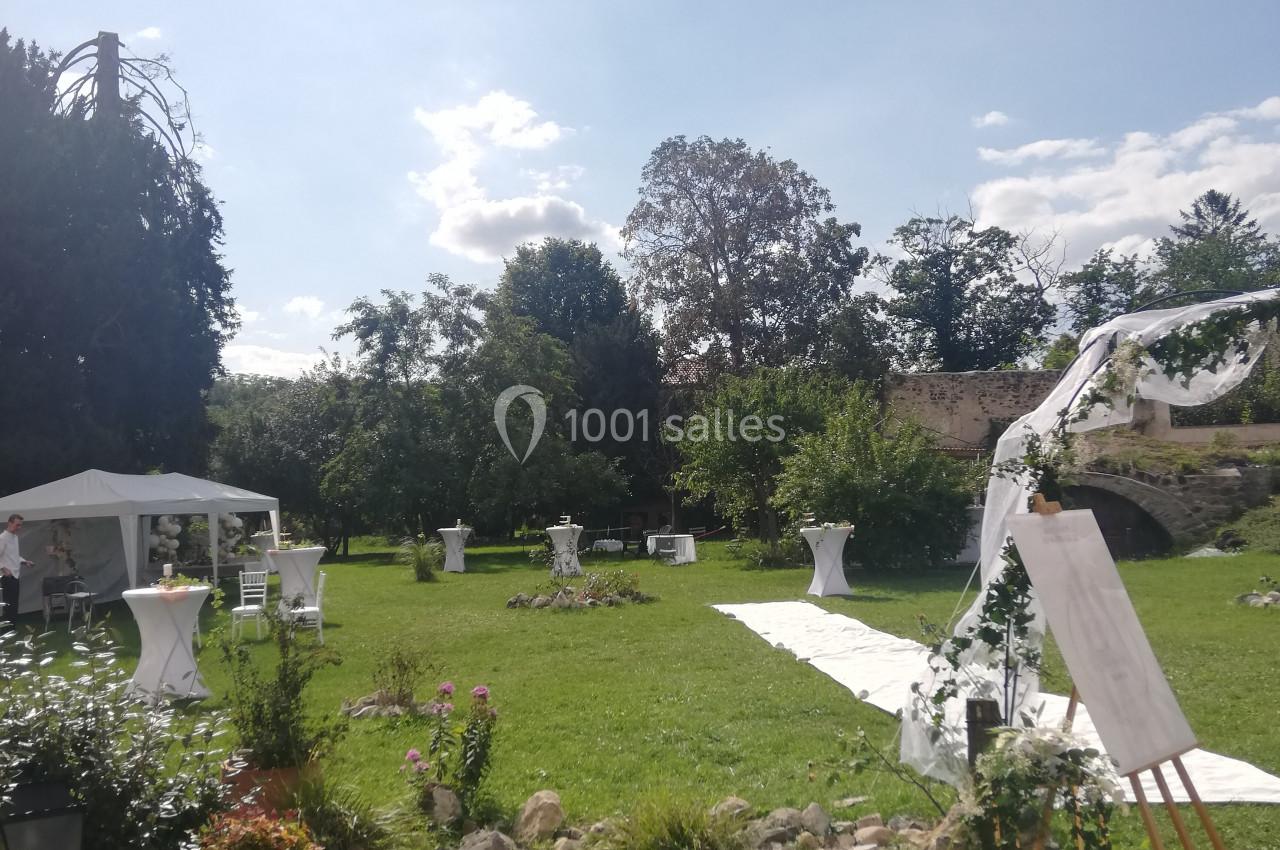 Jardin ensoleillé aménagé pour un événement avec tables hautes, tente blanche et allée décorée de fleurs.