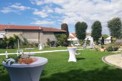 Jardin ensoleillé aménagé pour un événement avec tables hautes, tente blanche et allée décorée de fleurs.
