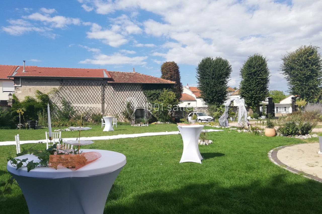 Jardin aménagé pour un événement avec tables hautes, arche décorative et ciel partiellement nuageux.