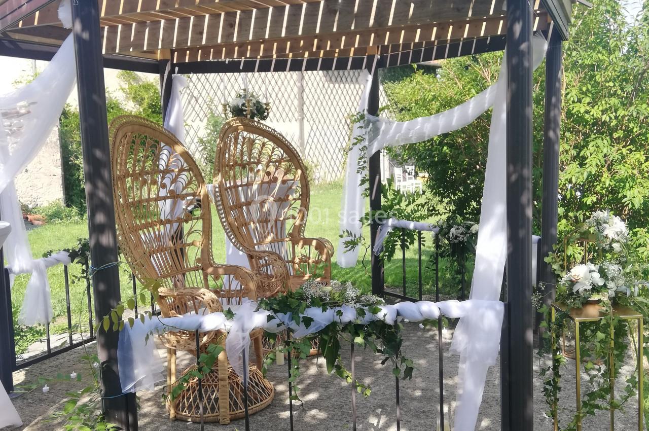 Tonnelle décorée de voilages blancs et de fleurs, avec des fauteuils en rotin, dans un jardin ensoleillé.