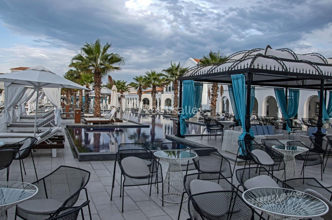 Terrasse d'un hôtel avec piscine, chaises, tables, lits balinais, palmiers et ciel nuageux en arrière-plan.