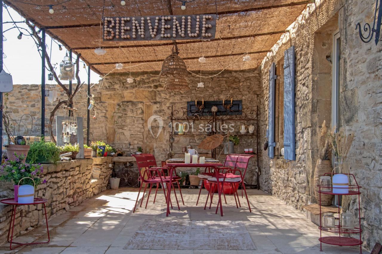 Terrasse en pierre avec mobilier rouge, décor rustique et enseigne ’Bienvenue’ sous une pergola ombragée.