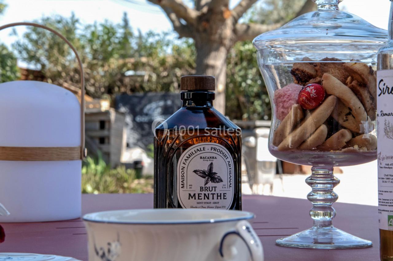 Bouteille de sirop de menthe posée sur une table avec des biscuits dans une bonbonnière et un fond extérieur ensoleillé.