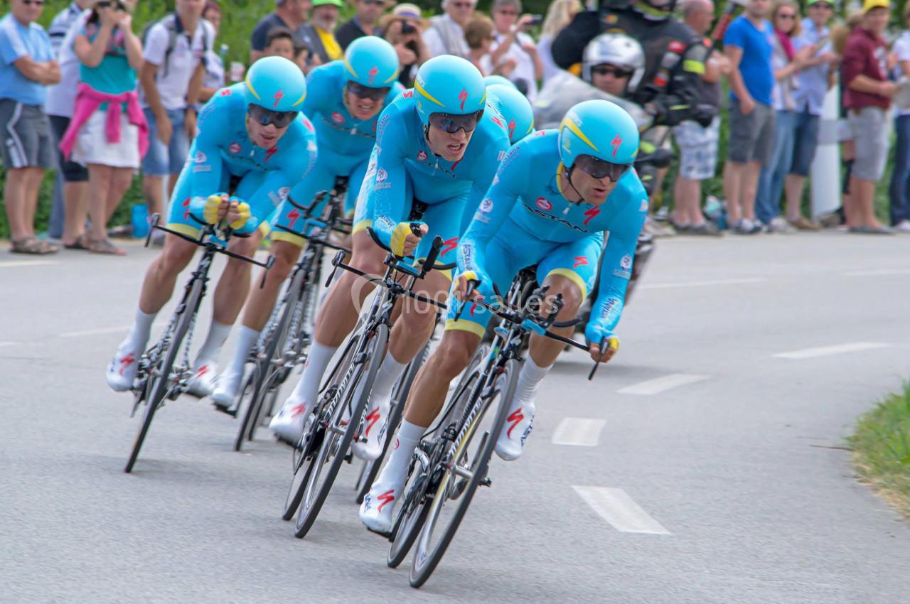 Quatre cyclistes en tenue bleue participent à une course en contre-la-montre, entourés de spectateurs au bord de la route.