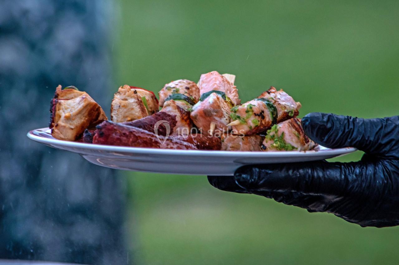 Assiette tenue par une main gantée, contenant des morceaux de viande grillée et des brochettes garnies d'herbes.