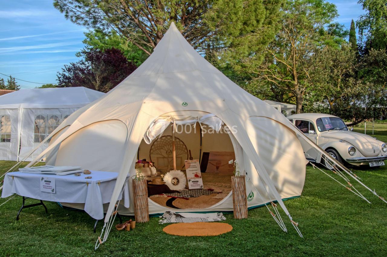 Tente beige aménagée avec des décorations bohèmes, installée sur une pelouse près d'une voiture vintage et d'arbres.