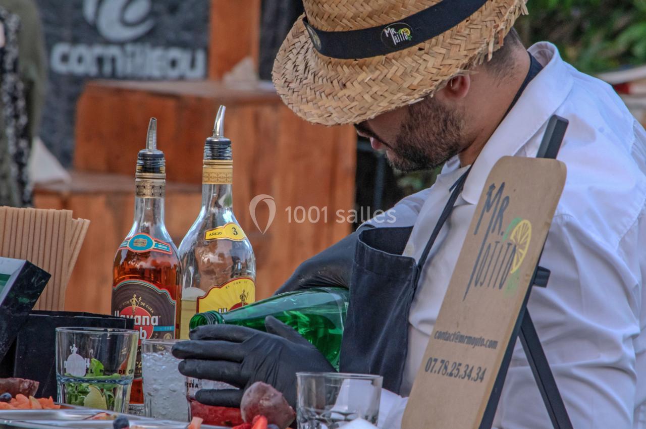 Un barman portant un chapeau prépare un cocktail avec des bouteilles d'alcool et des ingrédients frais sur un comptoir.