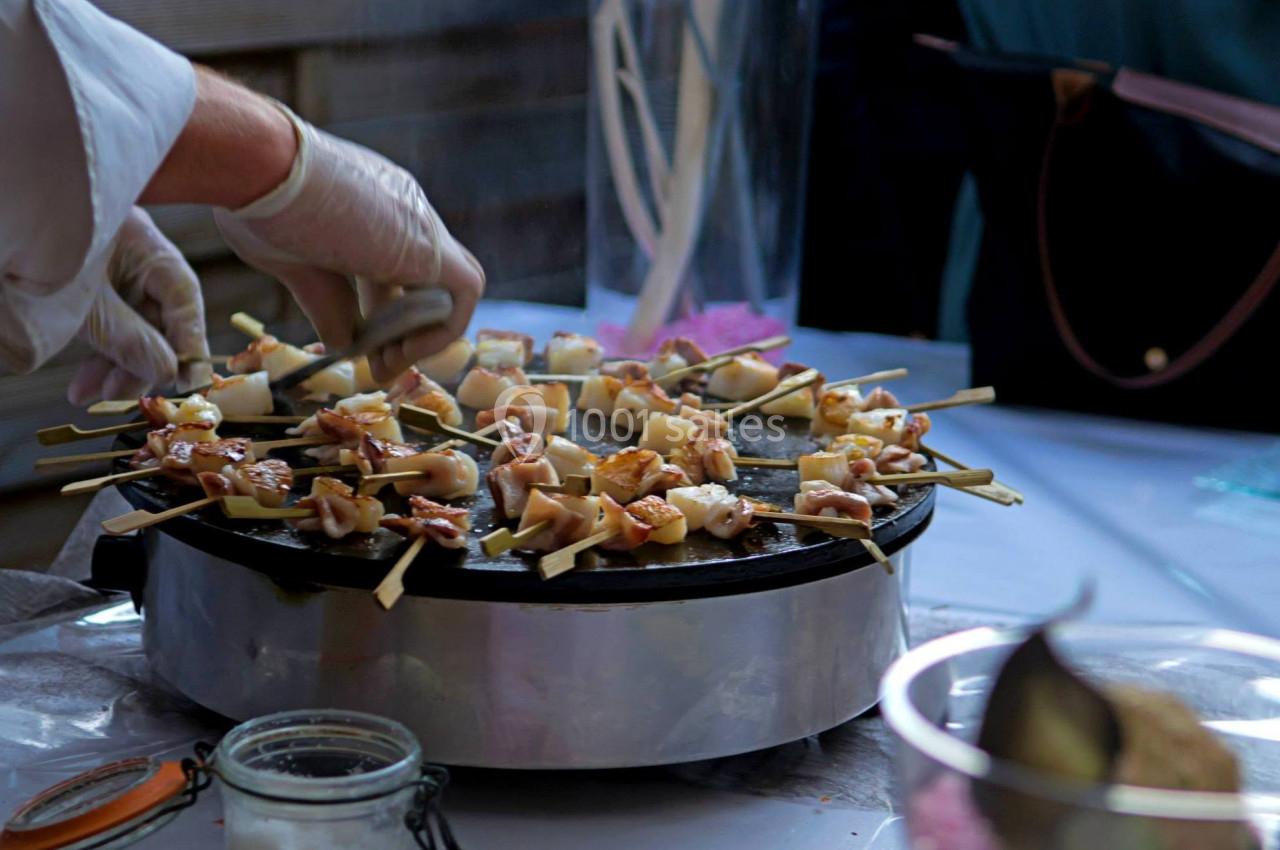 Des brochettes de viande et légumes sont grillées sur une plaque chauffante lors d'une préparation culinaire en extérieur.