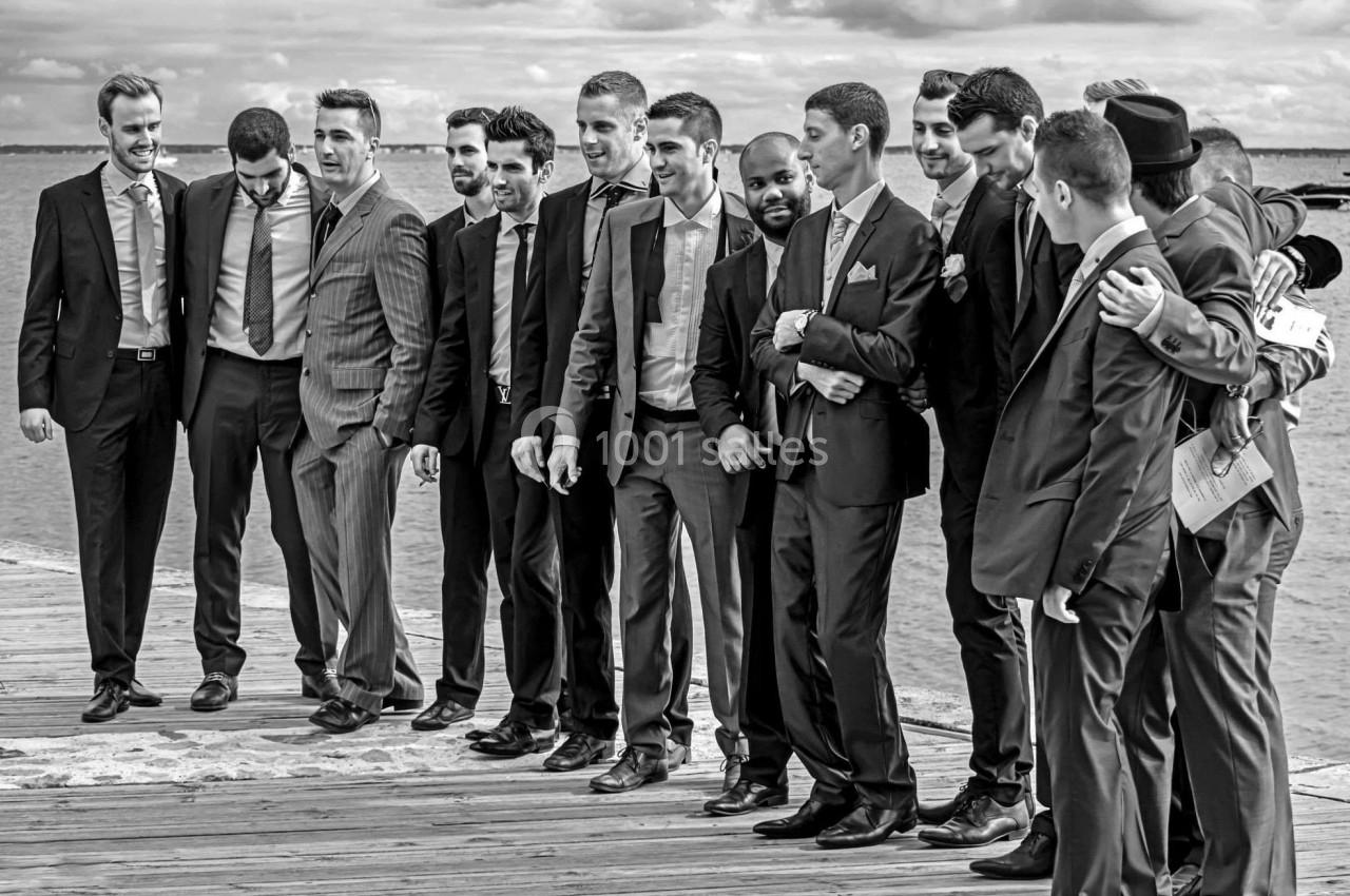 Un groupe d'hommes en costume posant sur un pont en bois près d'un plan d'eau sous un ciel nuageux.