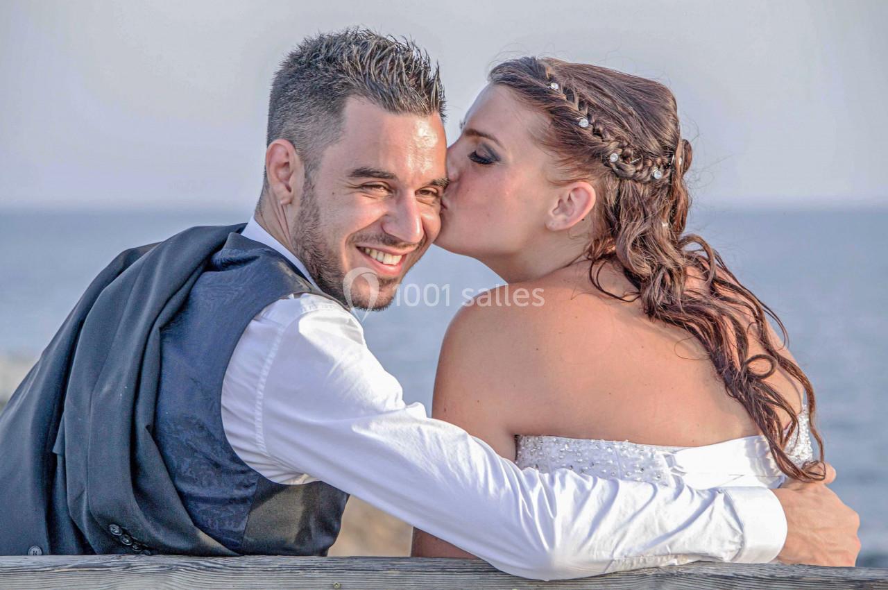 Un couple en tenue de mariage, assis près de la mer, la mariée embrassant le marié sur la joue.