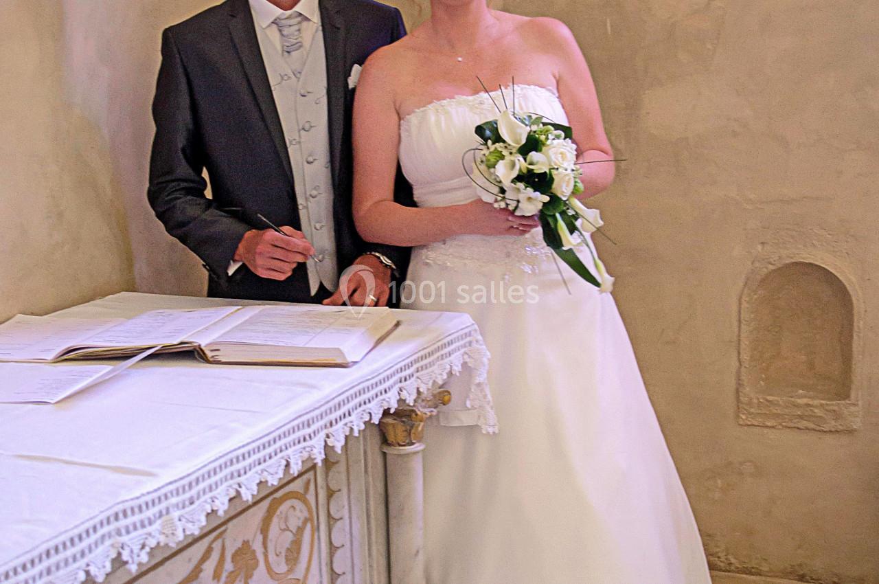 Un couple en tenue de mariage pose devant un autel décoré dans une pièce aux murs clairs.