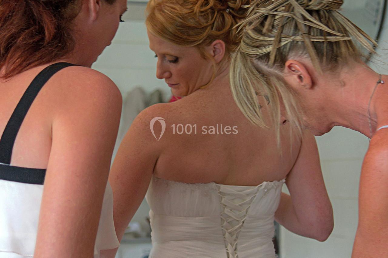 Deux femmes aident une mariée à ajuster le laçage de sa robe blanche dans une pièce lumineuse.