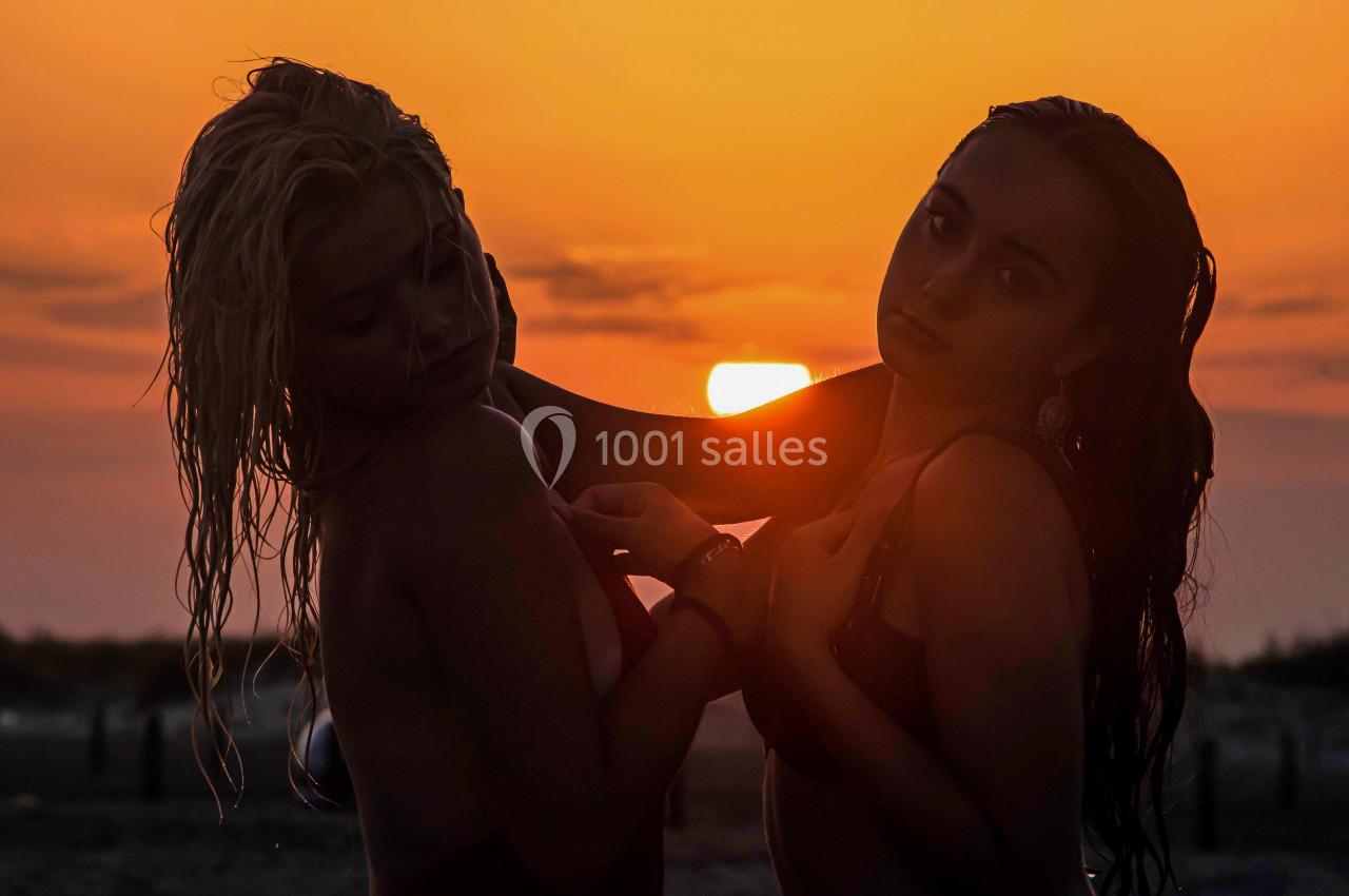 Deux femmes en maillot de bain posent devant un coucher de soleil, avec des cheveux mouillés et un arrière-plan sombre.