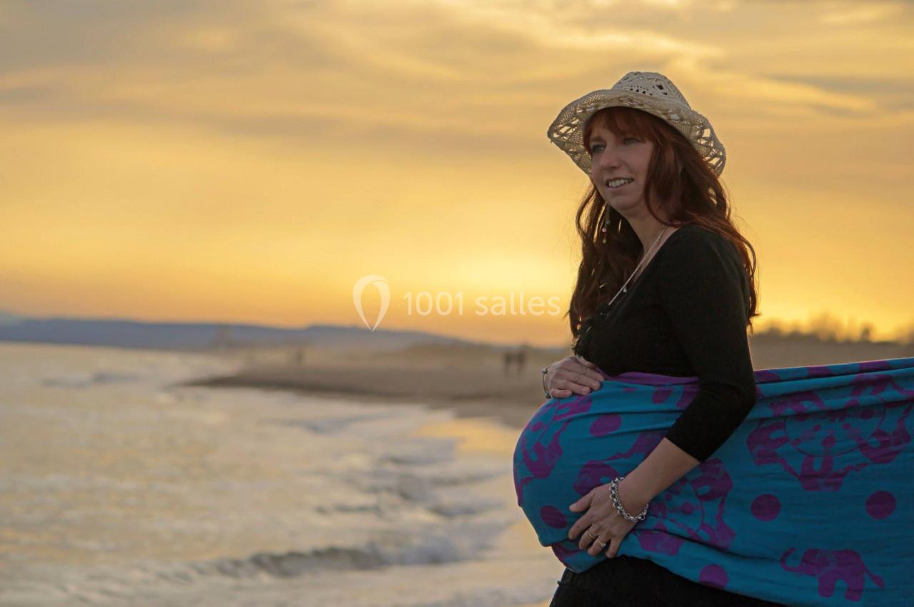Femme enceinte souriante portant un chapeau et un tissu coloré, debout sur une plage au coucher du soleil.