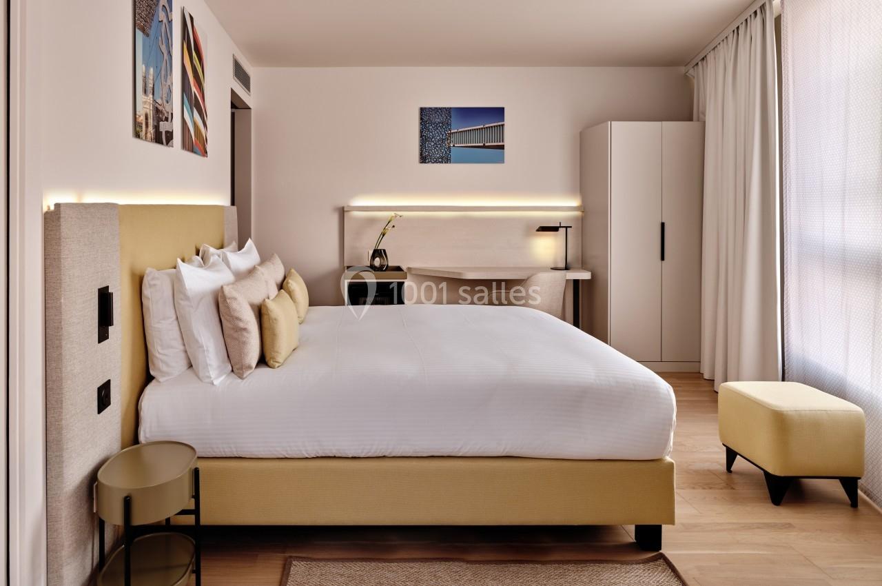 Chambre moderne avec lit double, bureau, éclairage doux, décor minimaliste et grande fenêtre avec rideaux.