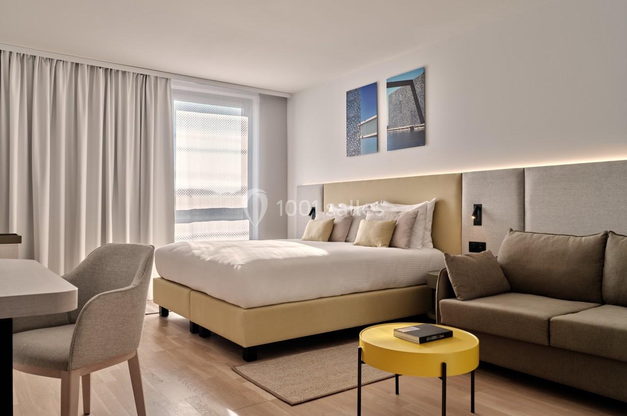 Chambre moderne avec lit double, canapé, table basse jaune et grande fenêtre avec rideaux blancs.