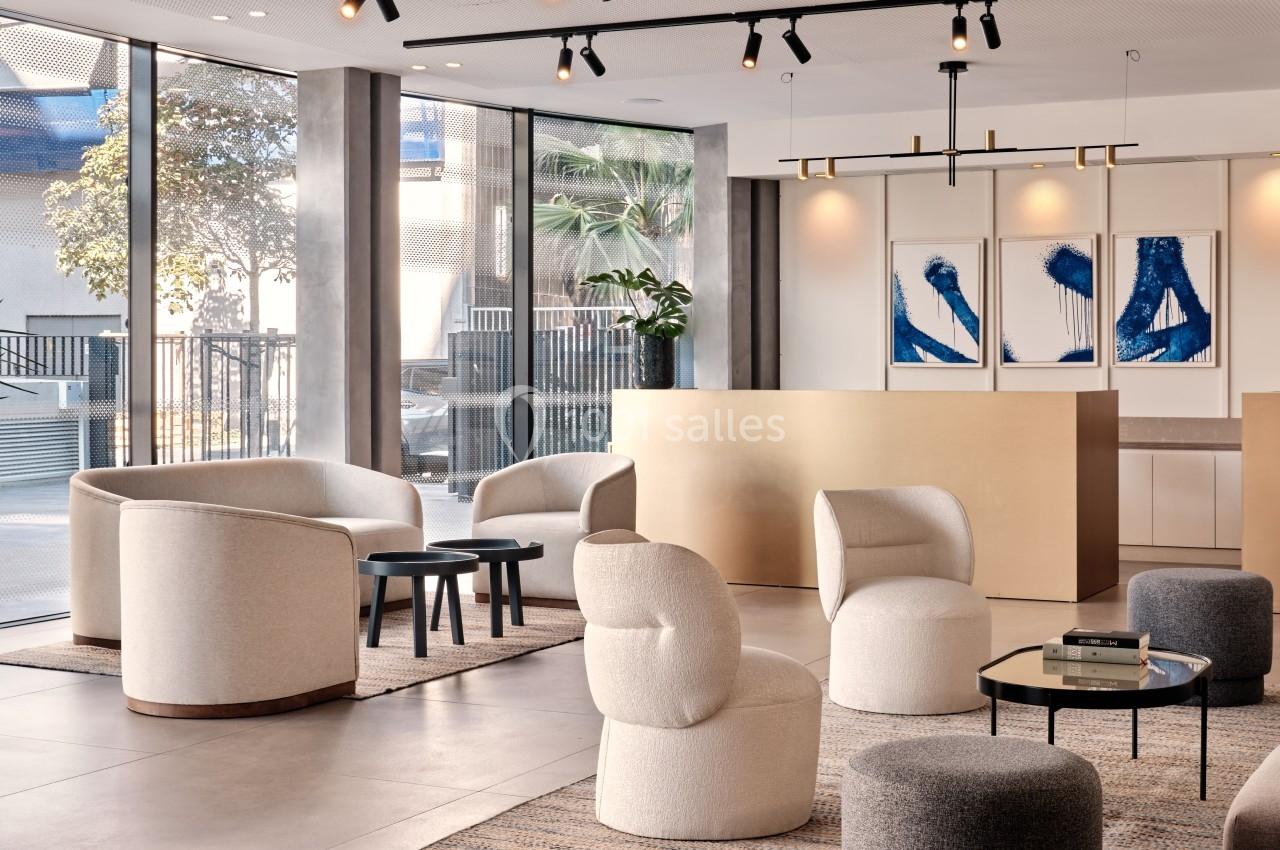 Espace lumineux avec fauteuils modernes, tables basses et comptoir beige, décoré de tableaux abstraits bleus.