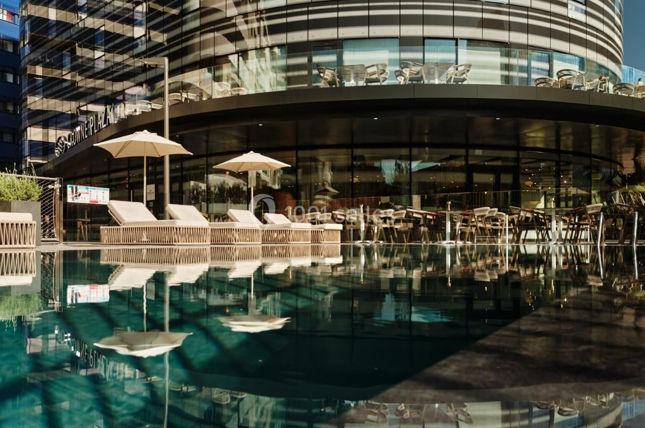 Façade d'un bâtiment moderne avec des baies vitrées, reflétée dans une piscine au premier plan.
