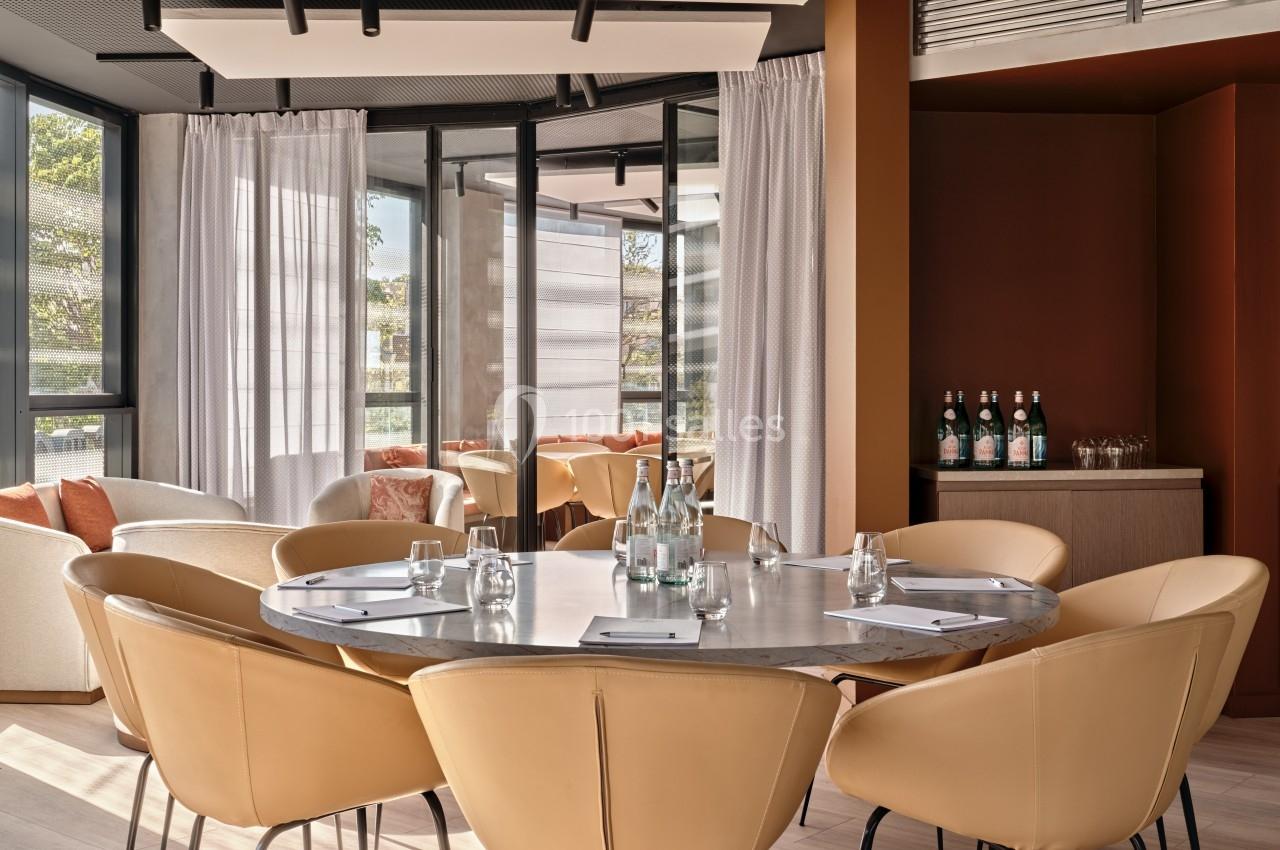 Salle de réunion lumineuse avec table ronde, chaises en cuir beige, baies vitrées et bouteilles d'eau sur un buffet.
