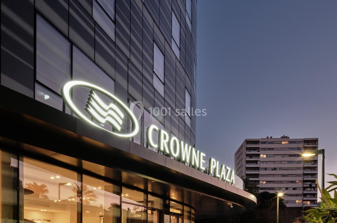 Façade d'un hôtel moderne avec enseigne lumineuse ’Crowne Plaza’ au crépuscule, entourée de végétation et d'immeubles.