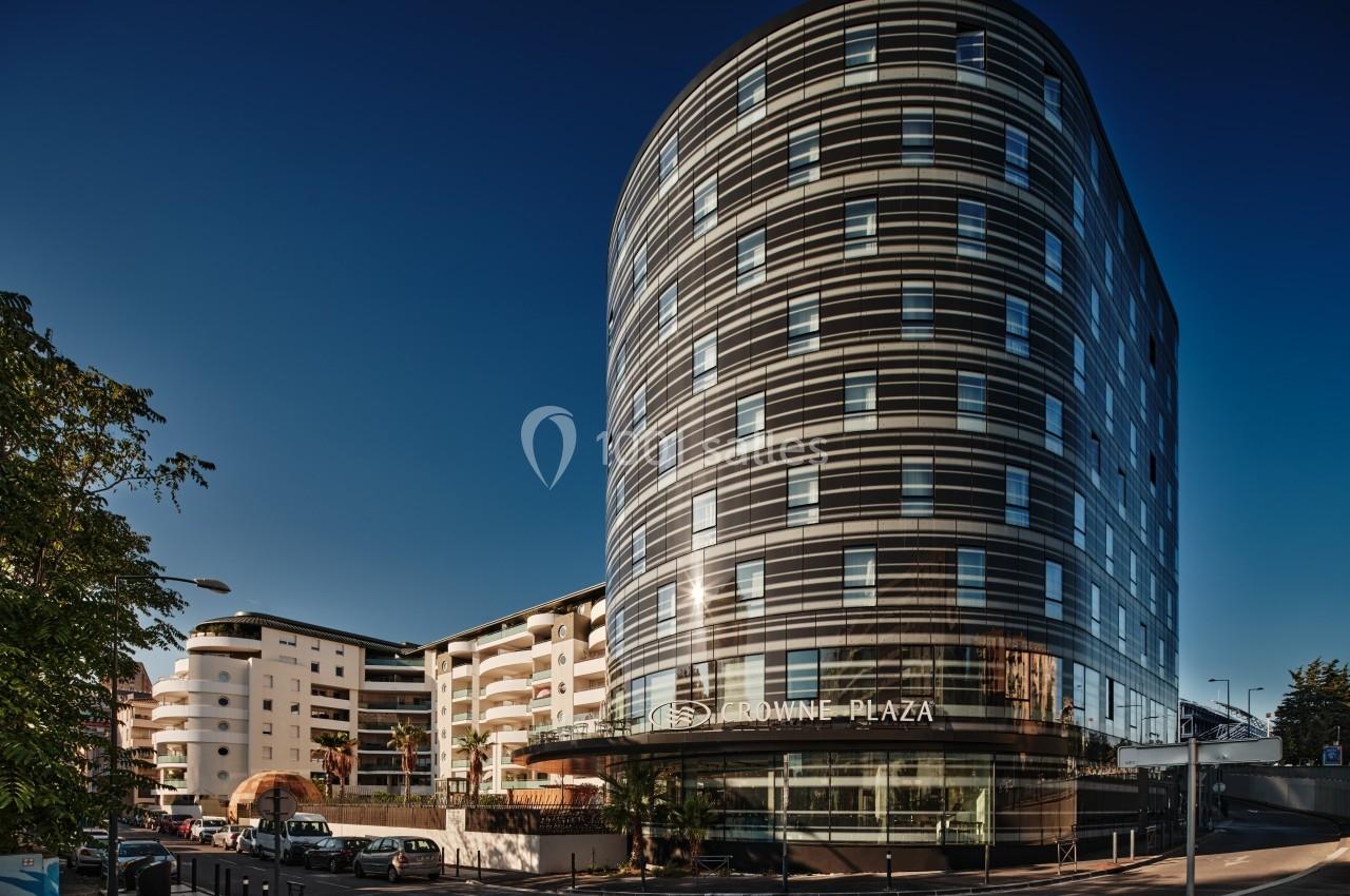 Bâtiment moderne en verre et métal, de forme arrondie, situé à un carrefour urbain sous un ciel dégagé.