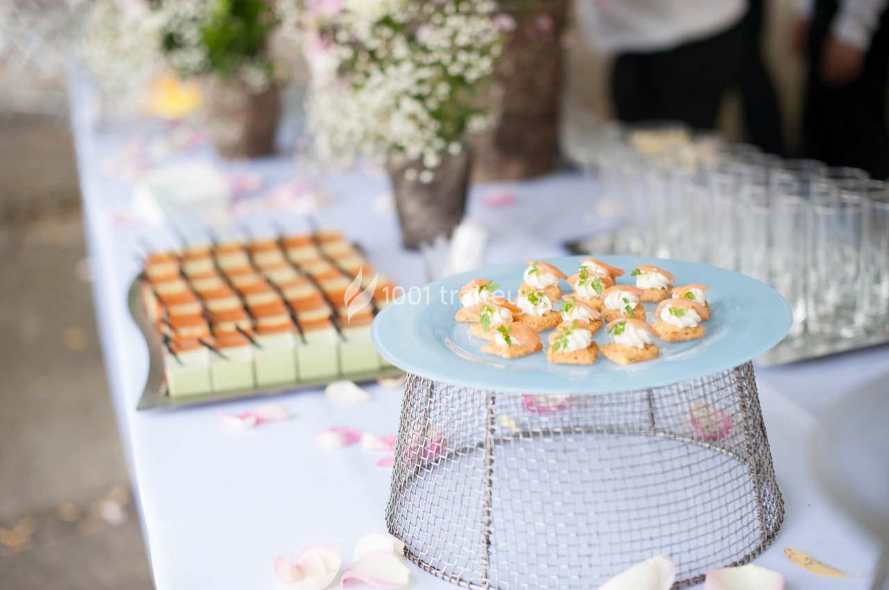 Plateau de bouchées apéritives sur une table décorée de fleurs, avec verrines et desserts en arrière-plan.