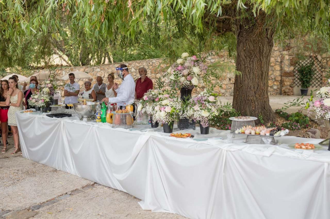 Buffet en extérieur sous des arbres, avec des invités se servant parmi des plats et boissons décorés de fleurs.