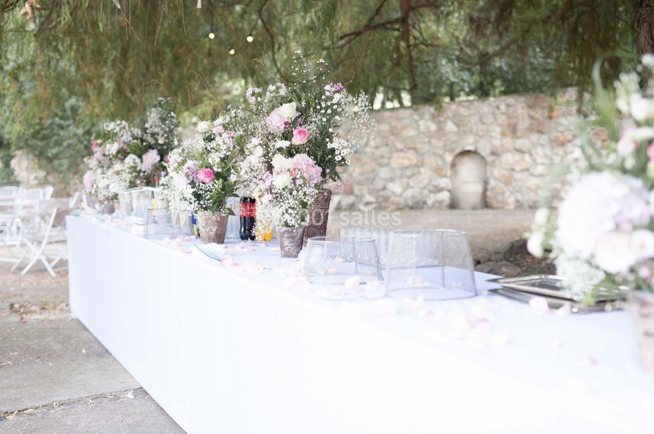 Table décorée pour un événement en extérieur, ornée de bouquets de fleurs roses et blanches sous des arbres.