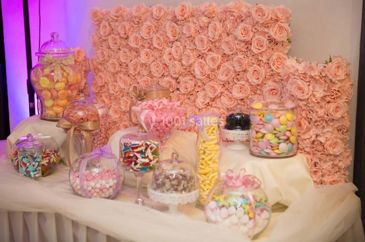 Buffet de bonbons colorés disposés dans des contenants en verre devant un mur décoré de roses roses.
