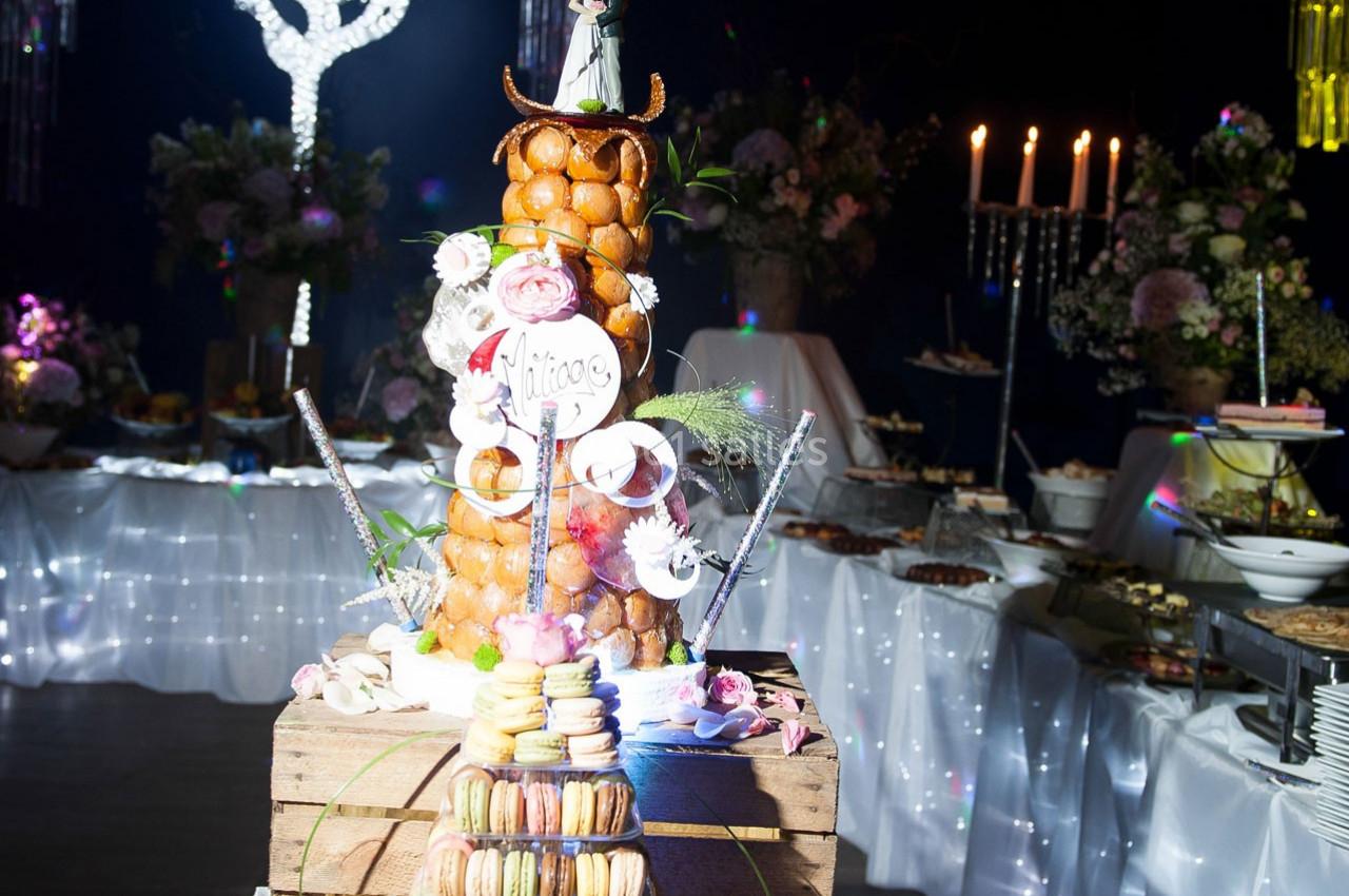 Pièce montée en choux et macarons décorée de fleurs, entourée de bougies et de plats sur une table de réception.