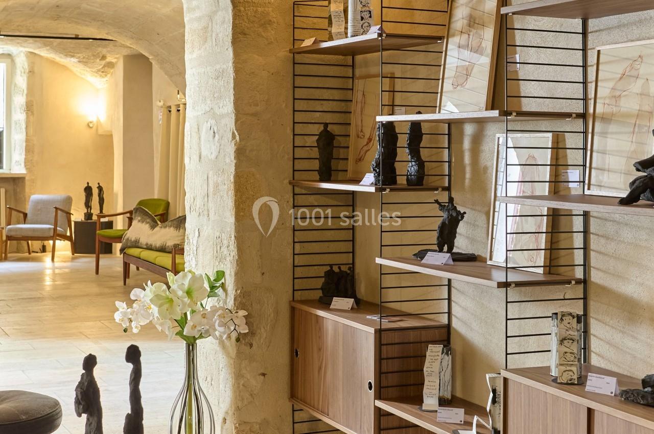 Étagères en bois et métal avec sculptures et dessins exposés dans une pièce en pierre lumineuse.