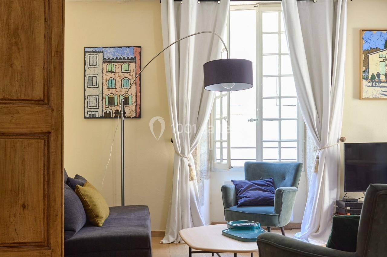 Salon lumineux avec parquet, canapé, fauteuils, table basse, lampadaire, rideaux blancs et fenêtre ouverte.