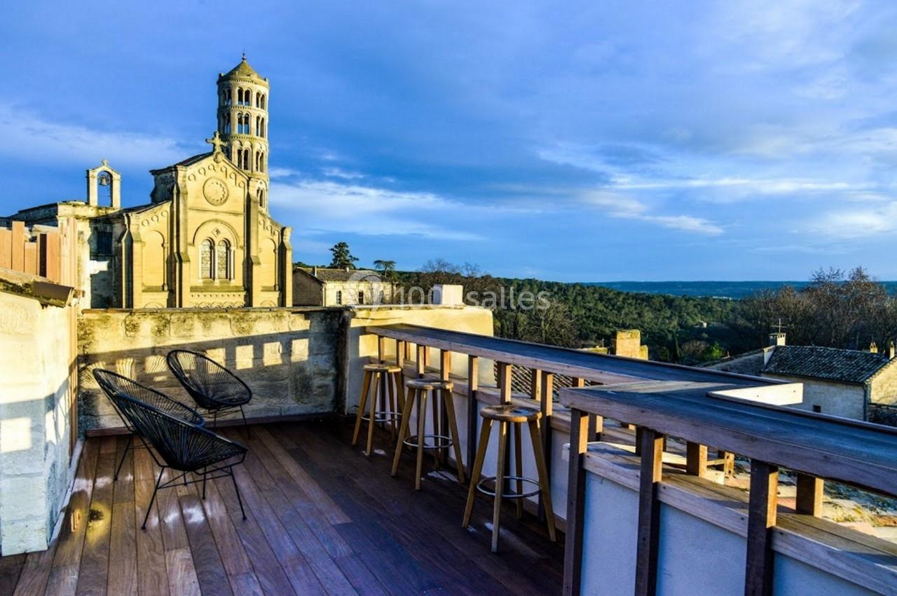 Terrasse en bois avec chaises et vue sur une église en pierre entourée de verdure sous un ciel partiellement nuageux.