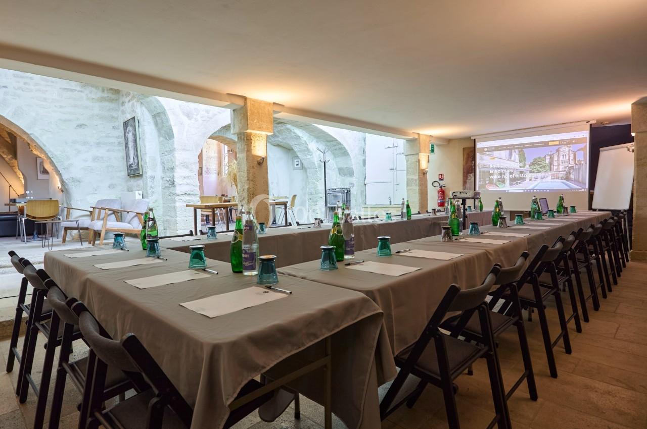 Salle de réunion avec tables disposées en U, chaises noires, bouteilles d'eau et écran de projection allumé.