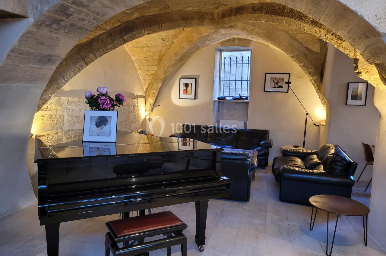 Salle voûtée en pierre avec un piano noir au premier plan, des fauteuils en cuir et des œuvres encadrées au mur.