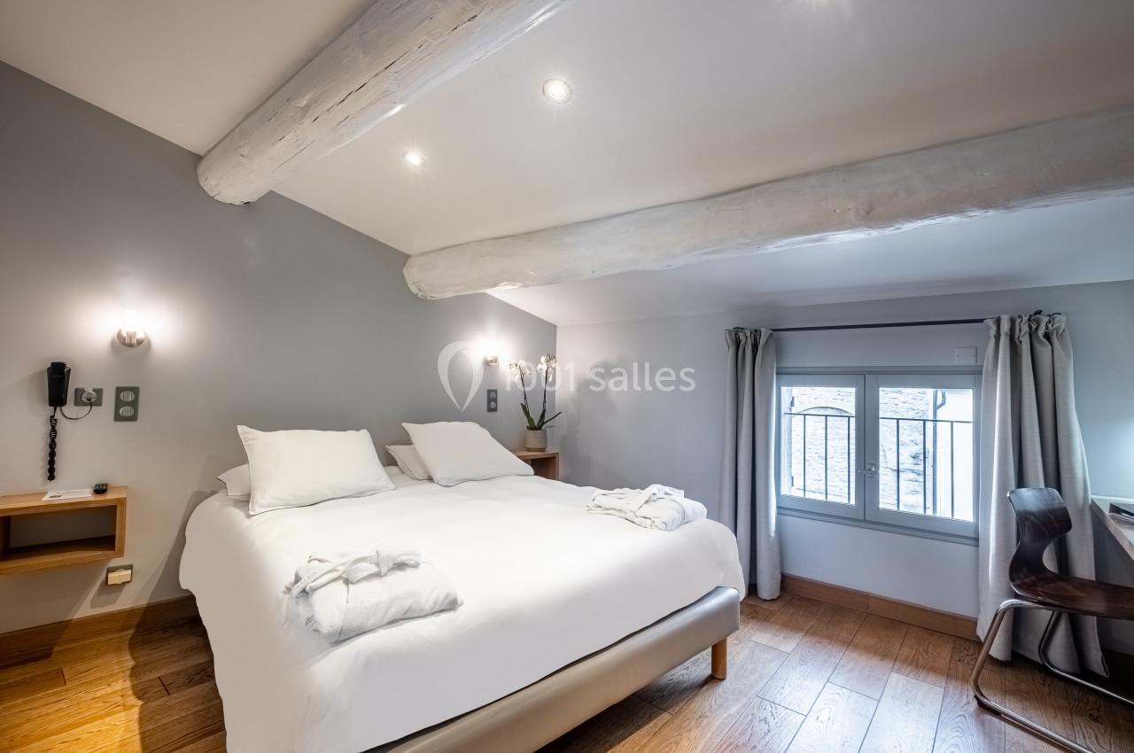 Chambre lumineuse avec lit double, poutres apparentes, parquet en bois, bureau et fenêtre avec rideaux gris.