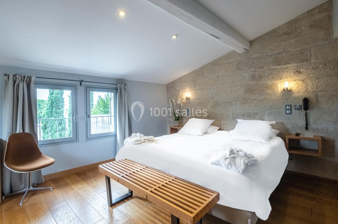 Chambre lumineuse avec lit double, murs en pierre, parquet, fenêtre donnant sur des arbres et mobilier minimaliste.