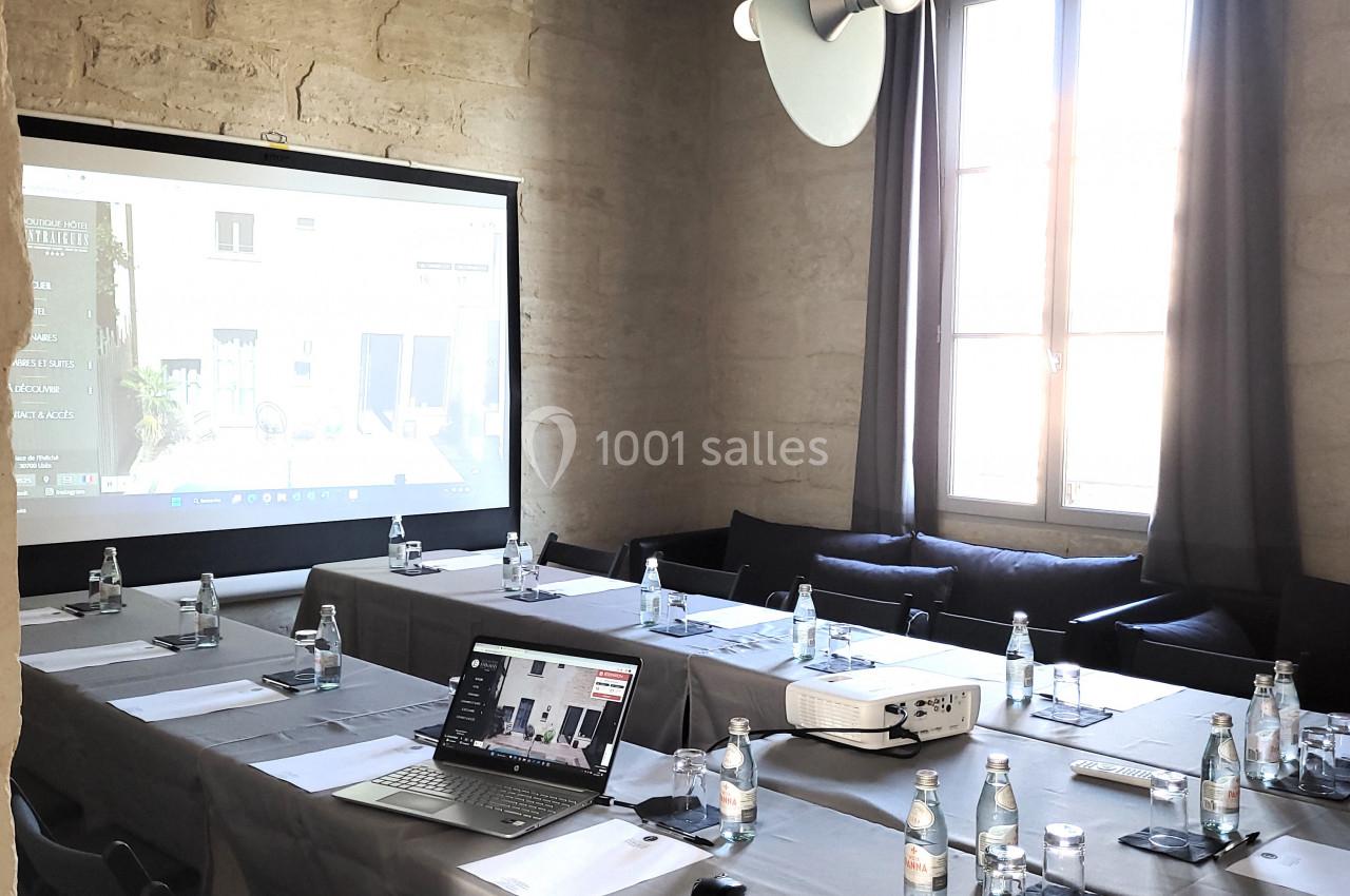 Salle de réunion lumineuse avec table rectangulaire, chaises noires, projecteur, écran et bouteilles d'eau.
