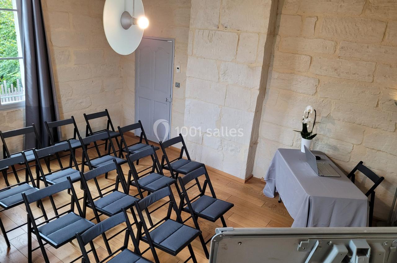 Salle lumineuse avec des chaises pliantes noires disposées devant une table recouverte d'une nappe blanche et un ordinateur…