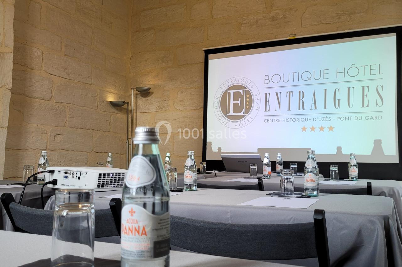 Salle de réunion avec tables dressées, bouteilles d'eau, verres, projecteur et écran affichant un logo d'hôtel.
