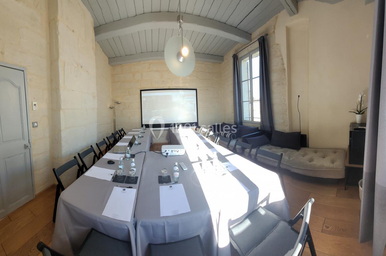 Salle de réunion lumineuse avec une grande table rectangulaire, chaises noires, écran de projection et murs en pierre claire.