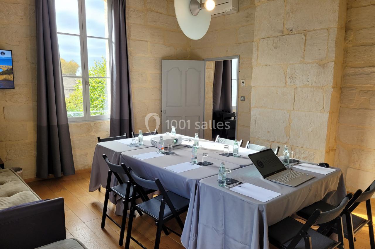 Salle de réunion lumineuse avec table rectangulaire dressée, chaises noires, murs en pierre et grande fenêtre donnant sur l…
